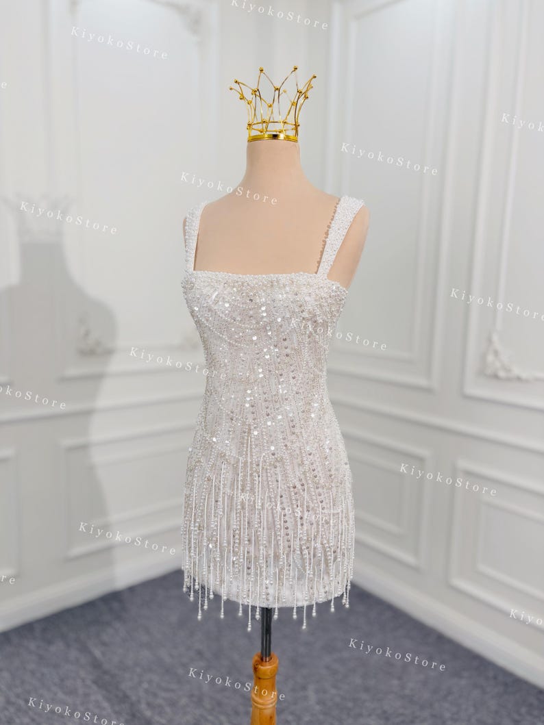 May include: A white, beaded mini dress with a square neckline and fringe detail. The dress is displayed on a mannequin with a gold crown. The dress is embellished with sequins and beads, creating a shimmering effect.
