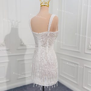 May include: A white, beaded mini dress on a mannequin, featuring a square neckline and beaded straps. The dress is adorned with sequins and dangling beads, and a gold crown sits atop the mannequin's head. The dress is displayed on a wooden stand.