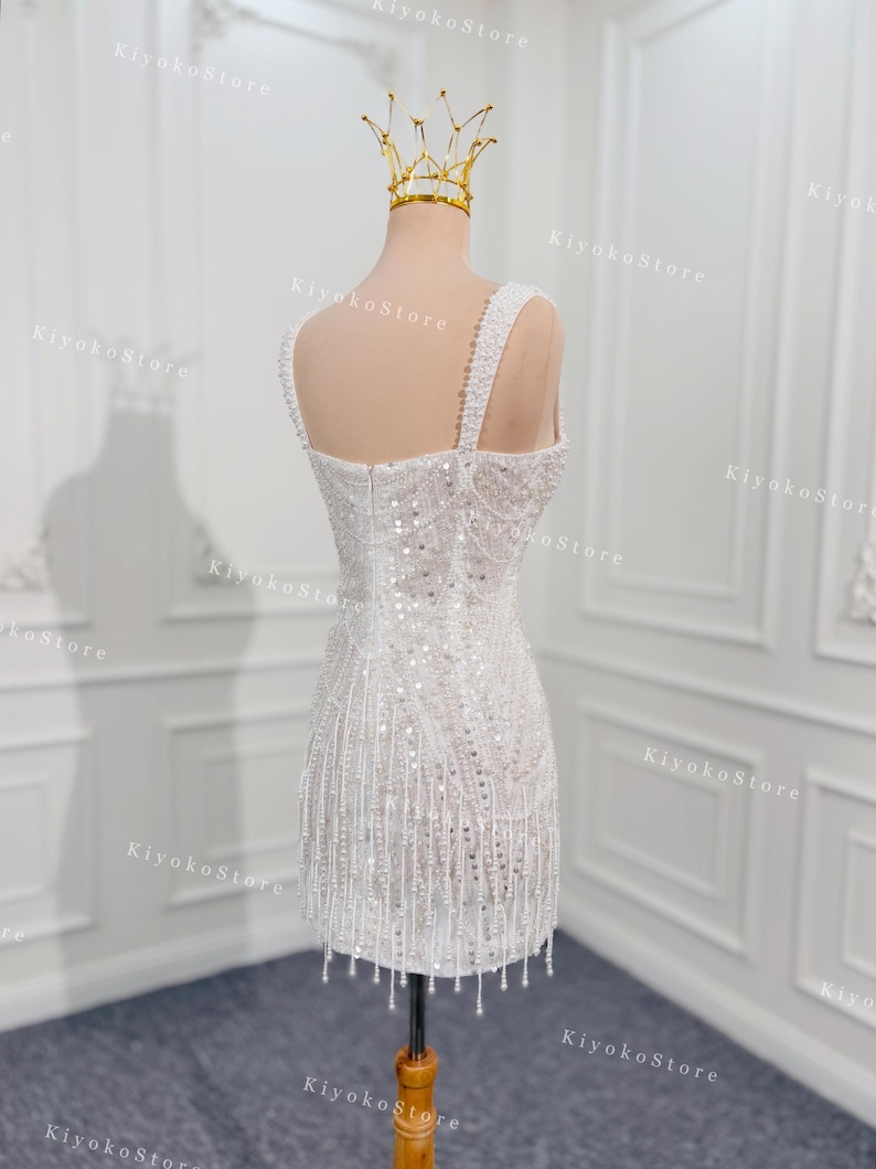 May include: A white, beaded mini dress on a mannequin, featuring a square neckline and beaded straps. The dress is adorned with sequins and dangling beads, and a gold crown sits atop the mannequin's head. The dress is displayed on a wooden stand.