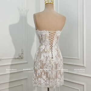 May include: A strapless white lace mini dress featuring a nude-colored lining, intricate floral embroidery, and a corset-style lace-up back closure. The dress is displayed on a mannequin wearing a gold wire crown, with KiyokoStore text visible.