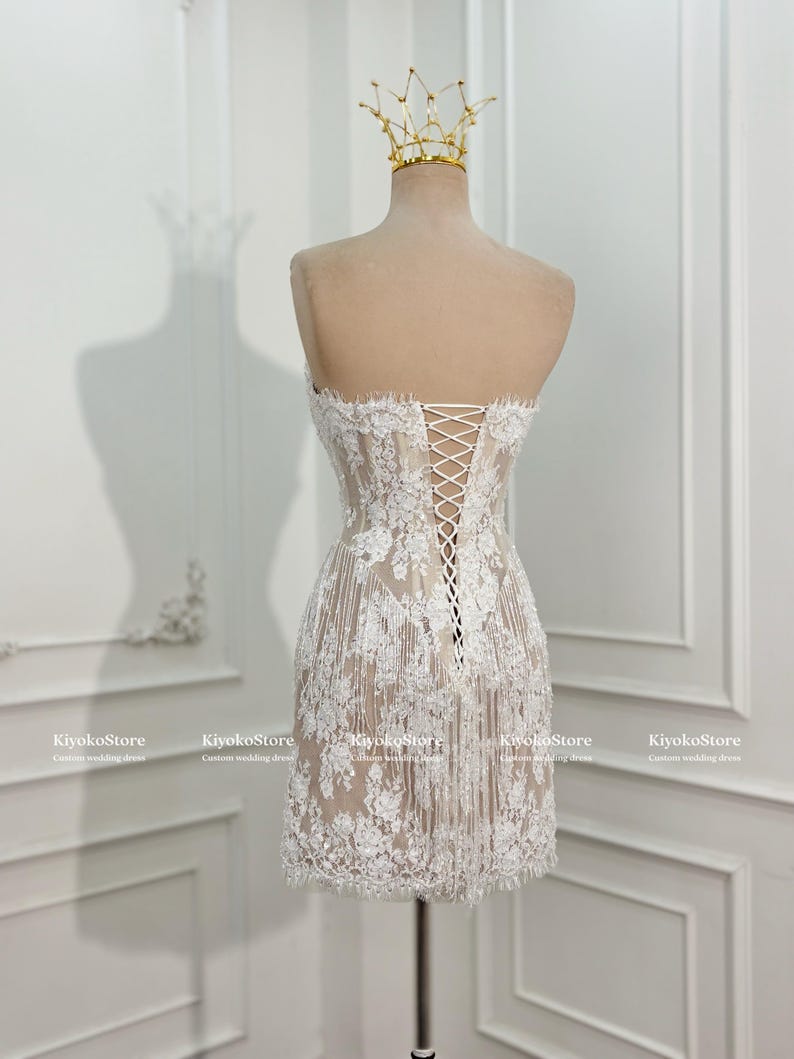 May include: A strapless white lace mini dress featuring a nude-colored lining, intricate floral embroidery, and a corset-style lace-up back closure. The dress is displayed on a mannequin wearing a gold wire crown, with KiyokoStore text visible.