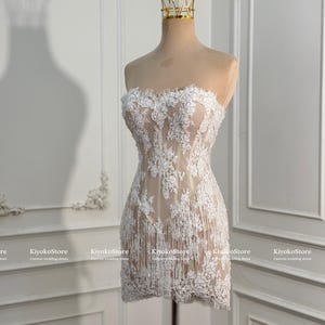 May include: A strapless mini dress featuring white floral lace embroidery over a nude-colored base. The garment includes a structured bodice and a skirt adorned with delicate white fringe detailing that hangs from the lace hemline.