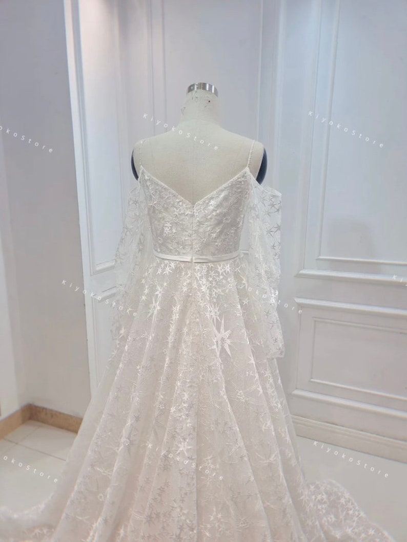 May include: A white wedding dress with a sweetheart neckline and a tulle skirt. The dress features a star-shaped pattern and a sheer overlay. The dress is shown from the back.