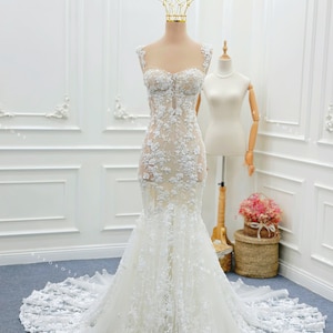 Pu&ograve; includere: Un abito da sposa a sirena bianco lungo fino a terra con scollo a cuore, spalline larghe e un intricato rivestimento in pizzo floreale su un corpetto trasparente. La gonna si allarga in un ampio strascico rifinito in pizzo.