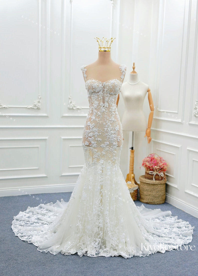 Pu&ograve; includere: Un abito da sposa a sirena bianco lungo fino a terra con scollo a cuore, spalline larghe e un intricato rivestimento in pizzo floreale su un corpetto trasparente. La gonna si allarga in un ampio strascico rifinito in pizzo.