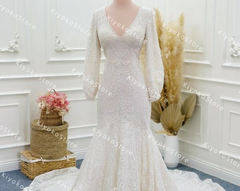 May include: A white, long-sleeved, beaded, mermaid-style wedding dress with a V-neckline. The dress has a fitted bodice and a flowing skirt.