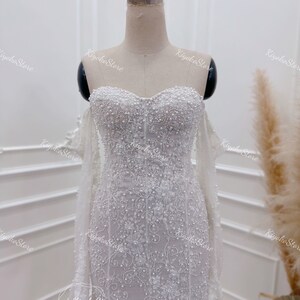May include: A white wedding dress with a sweetheart neckline and a fitted bodice. The dress is covered in delicate beading and lace, and has a long train.