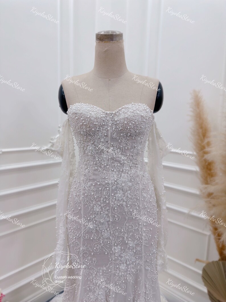 May include: A white wedding dress with a sweetheart neckline and a fitted bodice. The dress is covered in delicate beading and lace, and has a long train.