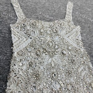 K&ouml;nnte beinhalten: Nahaufnahme eines wei&szlig;en, reich verzierten Kleides mit glitzernden Kristallen und Perlen. Das Kleid hat einen quadratischen Ausschnitt und d&uuml;nne Tr&auml;ger. Das Oberteil ist stark mit floralen und geometrischen Mustern verziert, was einen luxuri&ouml;sen und eleganten Look erzeugt.