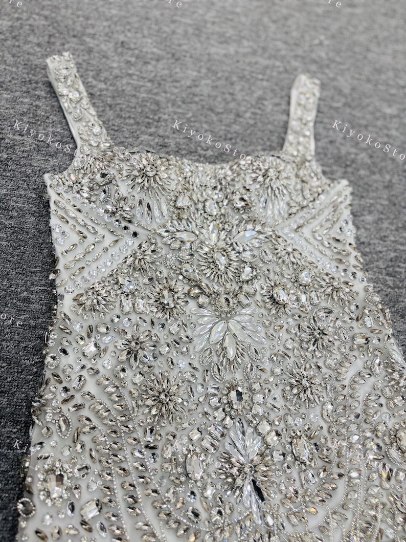 K&ouml;nnte beinhalten: Nahaufnahme eines wei&szlig;en, reich verzierten Kleides mit glitzernden Kristallen und Perlen. Das Kleid hat einen quadratischen Ausschnitt und d&uuml;nne Tr&auml;ger. Das Oberteil ist stark mit floralen und geometrischen Mustern verziert, was einen luxuri&ouml;sen und eleganten Look erzeugt.