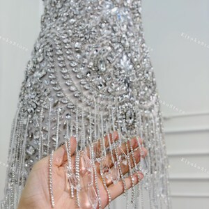 K&ouml;nnte beinhalten: Silbernes, stark verziertes Kleid mit aufwendiger Perlenarbeit und einem Saum aus h&auml;ngenden Kristallakzenten. Das Kleid ist &auml;rmellos und scheint aus einem schimmernden Stoff gefertigt zu sein, perfekt f&uuml;r einen besonderen Anlass.
