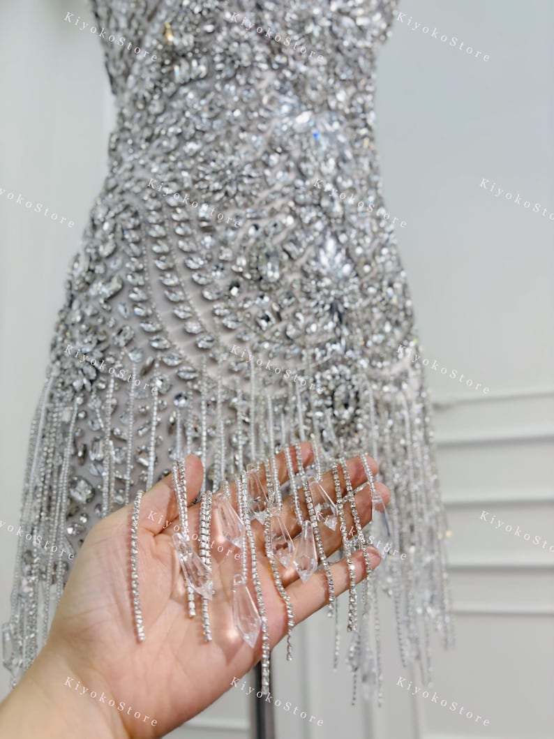 K&ouml;nnte beinhalten: Silbernes, stark verziertes Kleid mit aufwendiger Perlenarbeit und einem Saum aus h&auml;ngenden Kristallakzenten. Das Kleid ist &auml;rmellos und scheint aus einem schimmernden Stoff gefertigt zu sein, perfekt f&uuml;r einen besonderen Anlass.
