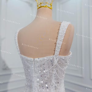 May include: A white wedding dress with a beaded bodice and straps, adorned with sequins. A gold crown sits atop a mannequin head. The dress is displayed on a mannequin in a well-lit room.