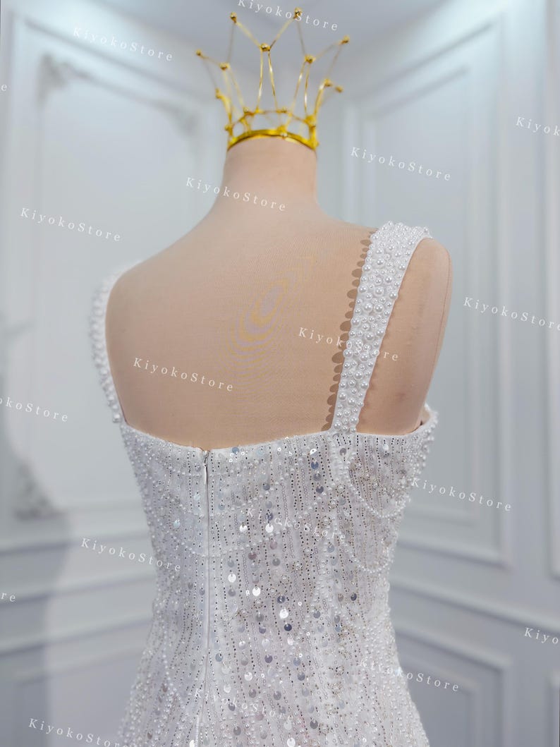 May include: A white wedding dress with a beaded bodice and straps, adorned with sequins. A gold crown sits atop a mannequin head. The dress is displayed on a mannequin in a well-lit room.