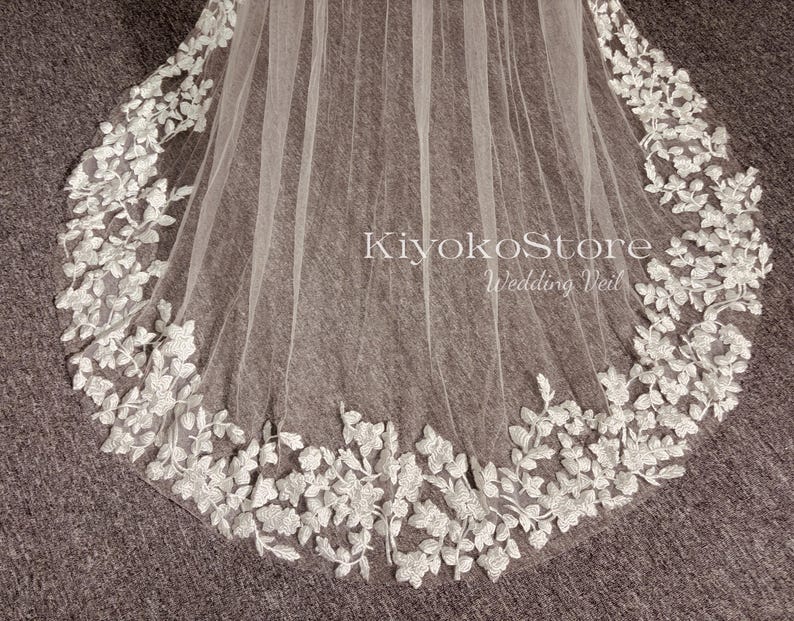 May include: A white wedding veil with a delicate floral lace trim. The lace features a repeating pattern of small leaves and flowers. The veil is made of soft tulle and is perfect for a bride looking for a romantic and elegant look.