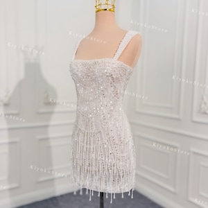 May include: A white, beaded mini dress with a square neckline and fringe detail. The dress is displayed on a mannequin and is topped with a gold crown. The dress has thick straps and is covered in sequins and beads.