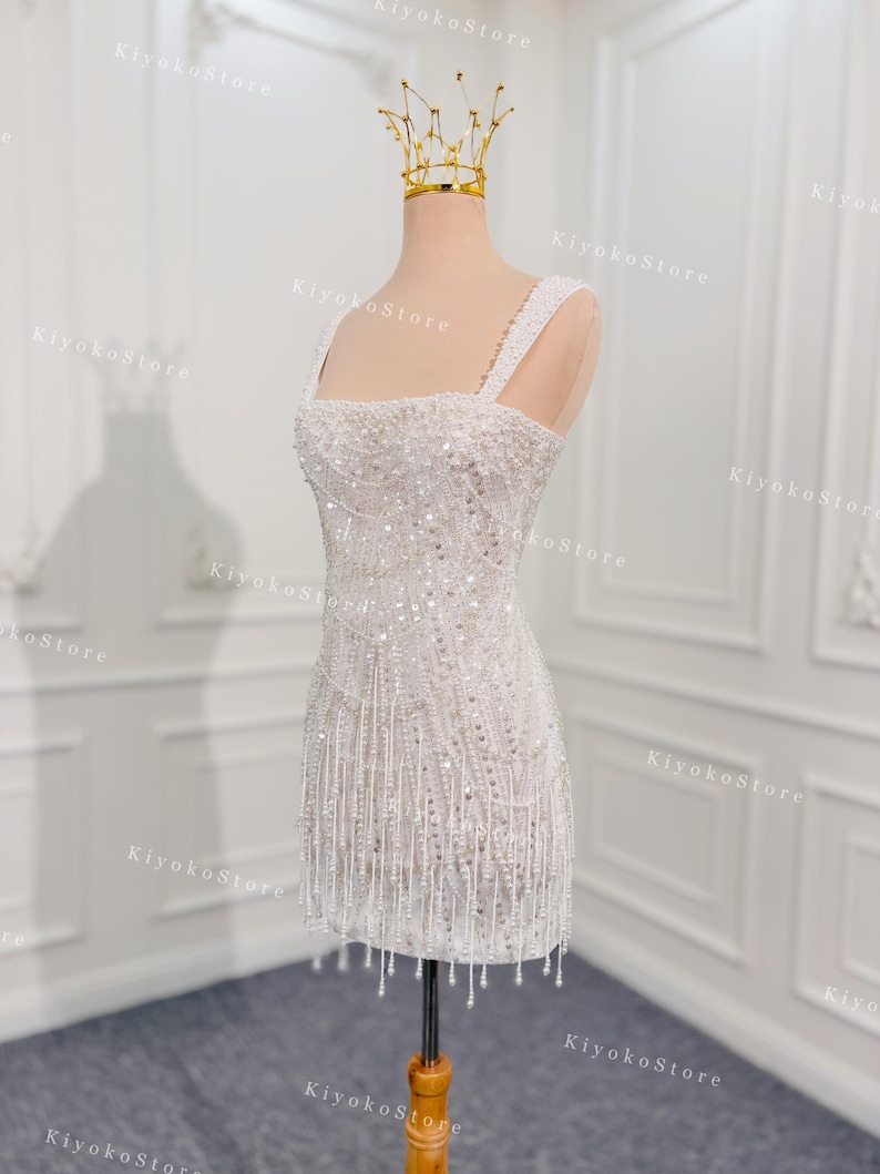 May include: A white, beaded mini dress with a square neckline and fringe detail. The dress is displayed on a mannequin and is topped with a gold crown. The dress has thick straps and is covered in sequins and beads.