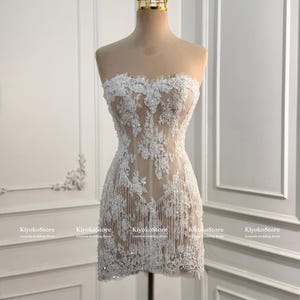 May include: A strapless mini dress featuring white floral lace embroidery over a nude-toned sheer base. The garment is adorned with scattered sequins and delicate fringe detailing along the hem. A gold-toned crown sits atop the mannequin.