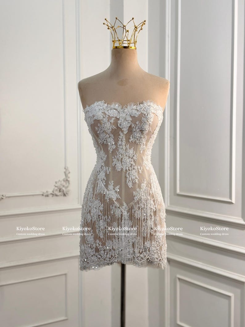 May include: A strapless mini dress featuring white floral lace embroidery over a nude-toned sheer base. The garment is adorned with scattered sequins and delicate fringe detailing along the hem. A gold-toned crown sits atop the mannequin.