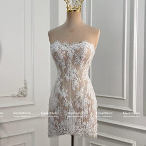 May include: A strapless mini dress featuring a nude-colored base with white floral lace overlays, intricate beadwork, and delicate fringe detailing along the hem. A gold-toned crown-shaped headpiece rests on top of the mannequin display.