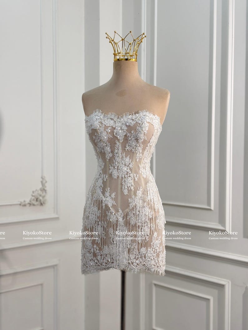 May include: A strapless mini dress featuring a nude-colored base with white floral lace overlays, intricate beadwork, and delicate fringe detailing along the hem. A gold-toned crown-shaped headpiece rests on top of the mannequin display.