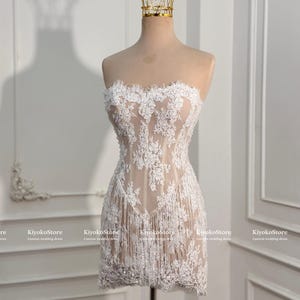 May include: A strapless mini dress featuring a nude-colored base with intricate white floral lace overlays and a fringe hem. The dress is displayed on a mannequin wearing a gold wire crown. The text KiyokoStore Custom wedding dress is visible.
