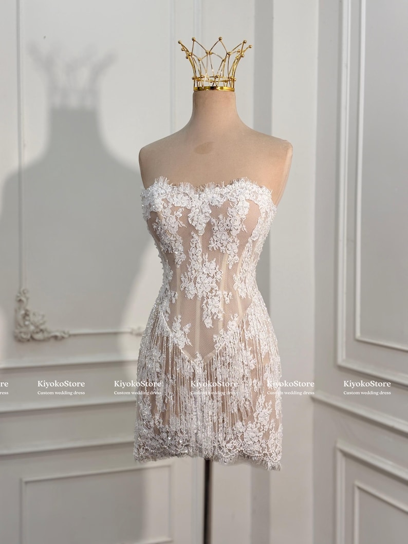 May include: A strapless mini dress featuring a nude-colored base with intricate white floral lace overlays and a fringe hem. The dress is displayed on a mannequin wearing a gold wire crown. The text KiyokoStore Custom wedding dress is visible.