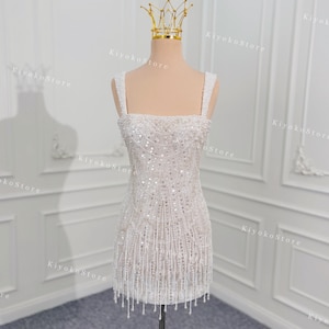 May include: A white, beaded mini dress with a square neckline and spaghetti straps. The dress is adorned with sequins and dangling beaded fringe. A gold crown sits atop a mannequin head. The dress is displayed on a wooden stand.