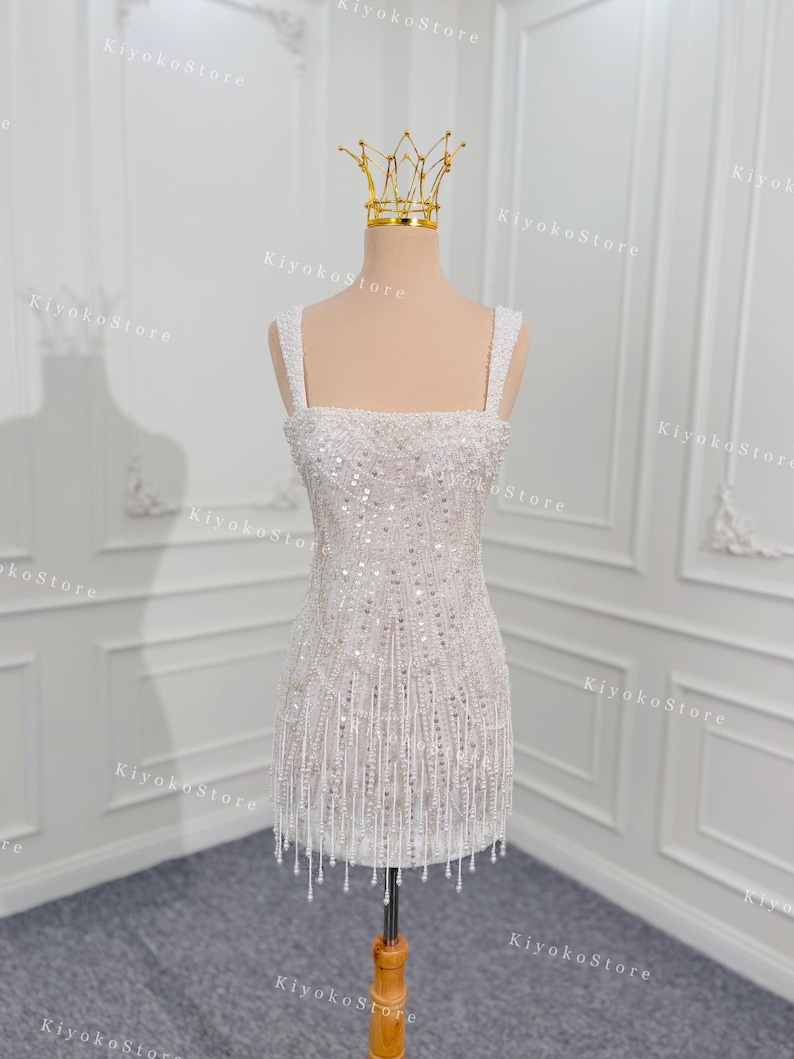 May include: A white, beaded mini dress with a square neckline and spaghetti straps. The dress is adorned with sequins and dangling beaded fringe. A gold crown sits atop a mannequin head. The dress is displayed on a wooden stand.