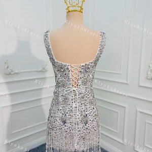 K&ouml;nnte beinhalten: Wei&szlig;es, verziertes Minikleid mit quadratischem Ausschnitt und Schn&uuml;rr&uuml;cken. Das Kleid ist mit silbernen Pailletten und Perlen besetzt und hat einen Perlenfransenbesatz am Saum. Eine goldene Krone sitzt auf dem Mannequinkopf.