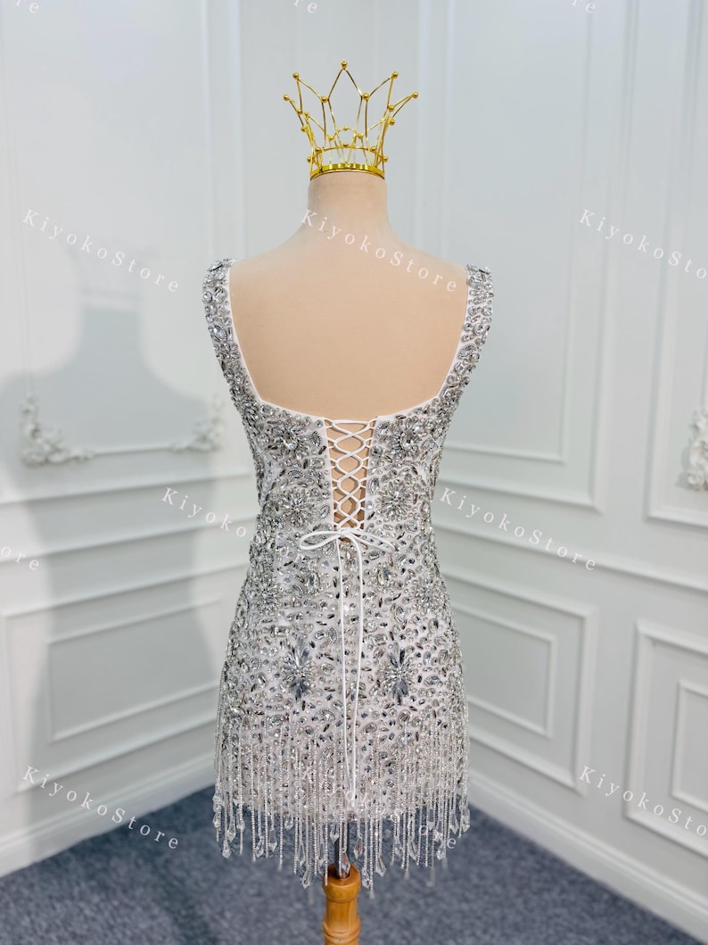 K&ouml;nnte beinhalten: Wei&szlig;es, verziertes Minikleid mit quadratischem Ausschnitt und Schn&uuml;rr&uuml;cken. Das Kleid ist mit silbernen Pailletten und Perlen besetzt und hat einen Perlenfransenbesatz am Saum. Eine goldene Krone sitzt auf dem Mannequinkopf.