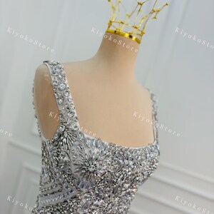 K&ouml;nnte beinhalten: Ein silbernes, verziertes Abendkleid mit quadratischem Ausschnitt und breiten Tr&auml;gern. Das Oberteil und die Tr&auml;ger sind reich mit glitzernden Kristallen und Strasssteinen verziert. Eine zarte goldene Krone sitzt auf dem Kopf der B&uuml;ste.
