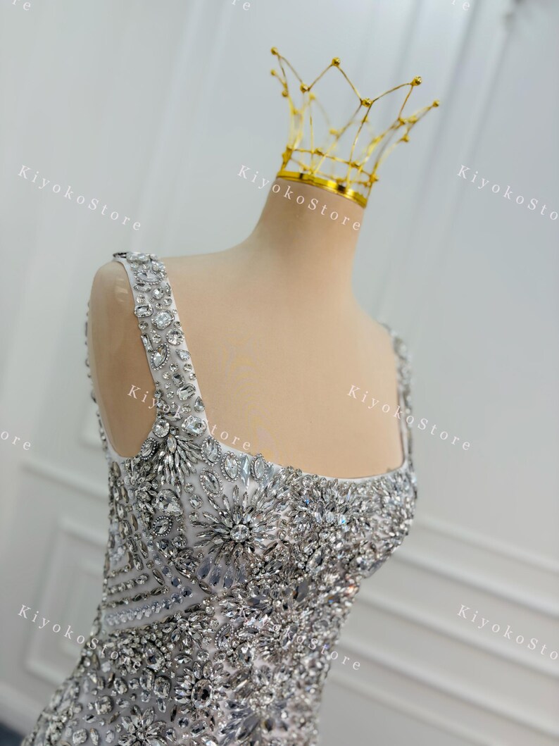 K&ouml;nnte beinhalten: Ein silbernes, verziertes Abendkleid mit quadratischem Ausschnitt und breiten Tr&auml;gern. Das Oberteil und die Tr&auml;ger sind reich mit glitzernden Kristallen und Strasssteinen verziert. Eine zarte goldene Krone sitzt auf dem Kopf der B&uuml;ste.
