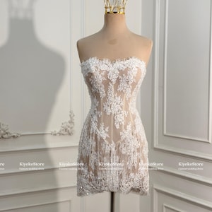 May include: A strapless mini dress featuring a nude-colored base layer with intricate white floral lace overlays and delicate fringe detailing along the hem. The garment is displayed on a mannequin topped with a gold-toned wire crown accessory.