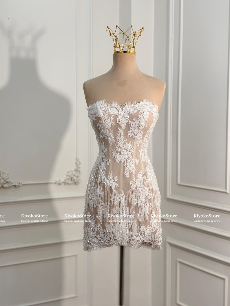 May include: A strapless mini dress featuring a nude-colored base layer with intricate white floral lace overlays and delicate fringe detailing along the hem. The garment is displayed on a mannequin topped with a gold-toned wire crown accessory.