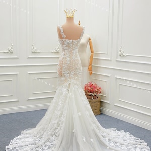 Pu&ograve; includere: Un abito da sposa bianco lungo fino a terra con un corpetto trasparente decorato da intricati ricami floreali in pizzo, spalline larghe e una silhouette aderente che si apre in un lungo strascico di tulle. L'abito &egrave; esposto su un manichino con una corona dorata.
