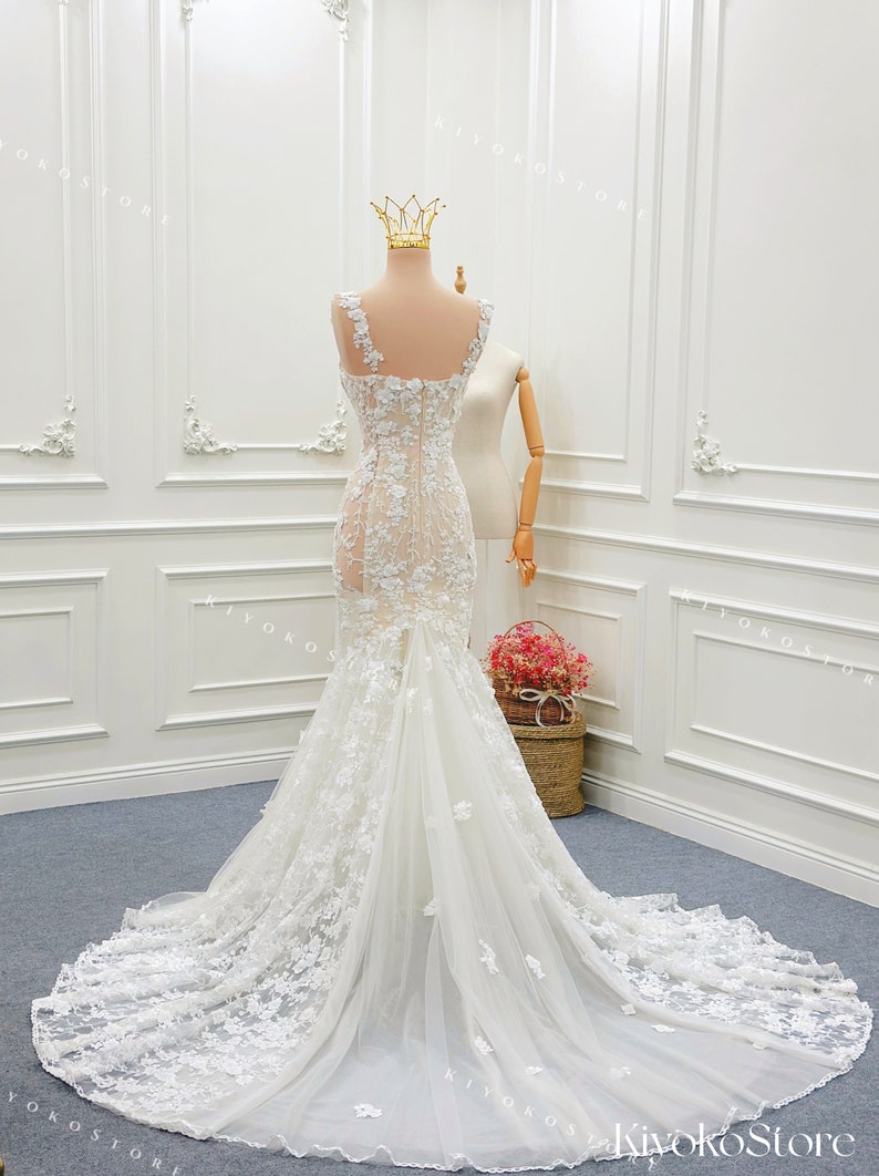 Pu&ograve; includere: Un abito da sposa bianco lungo fino a terra con un corpetto trasparente decorato da intricati ricami floreali in pizzo, spalline larghe e una silhouette aderente che si apre in un lungo strascico di tulle. L'abito &egrave; esposto su un manichino con una corona dorata.