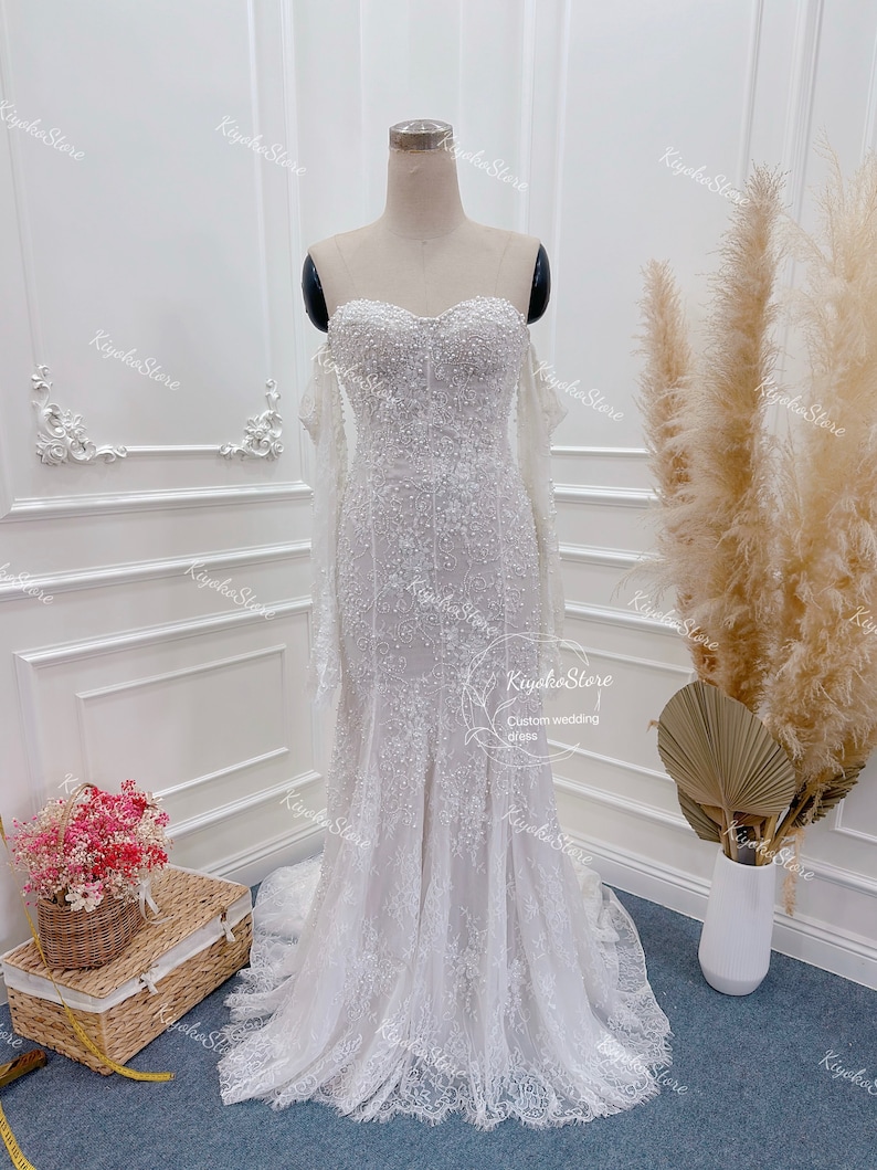 May include: A white off-the-shoulder wedding dress with a fitted bodice and a flowing skirt. The dress is covered in delicate beading and lace.