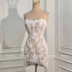 May include: A strapless mini dress featuring a nude-colored base with intricate white floral lace overlays and a fringe hemline. The dress is displayed on a mannequin wearing a gold wire crown. The text KiyokoStore Custom wedding dress is visible.