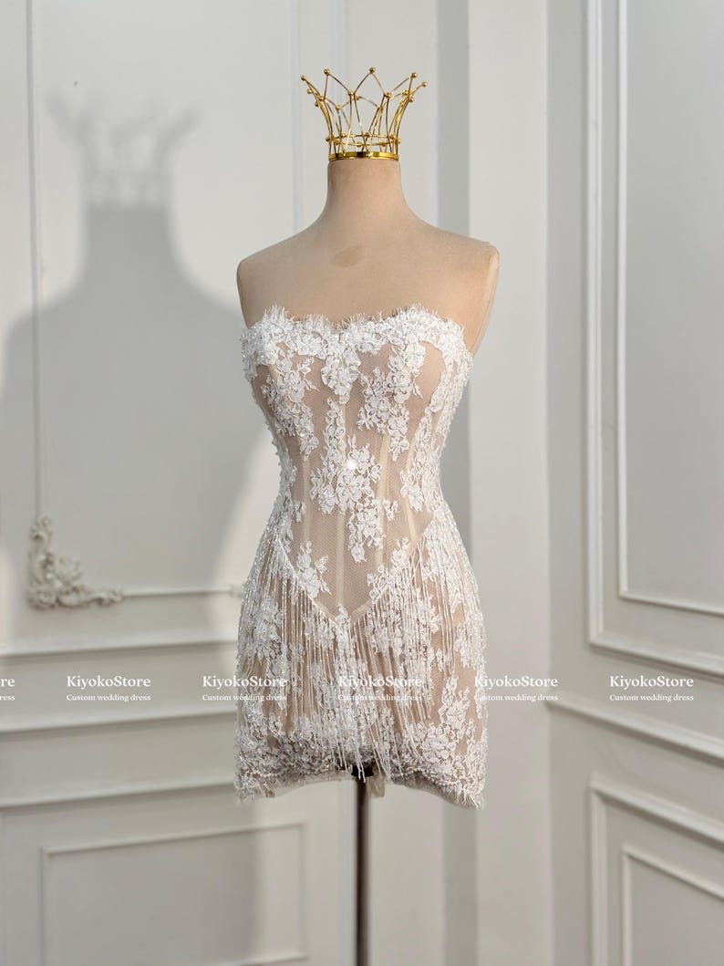 May include: A strapless mini dress featuring a nude-colored base with intricate white floral lace overlays and a fringe hemline. The dress is displayed on a mannequin wearing a gold wire crown. The text KiyokoStore Custom wedding dress is visible.