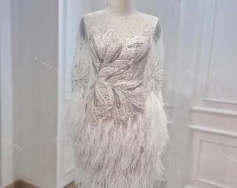 May include: A white, embellished mini dress with long sheer sleeves. The bodice features intricate silver beading and sequin detailing. The skirt is adorned with white feathers, creating a textured, elegant look. The dress is displayed on a mannequin.