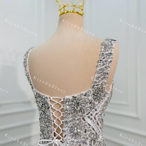 K&ouml;nnte beinhalten: Wei&szlig;es, verziertes Abendkleid mit glitzernden Kristall- und Strassdetails. Der R&uuml;cken hat einen Verschluss im Korsettstil und eine zarte Schleife. Eine goldene Krone sitzt auf einem Schaufensterpuppenkopf, was auf ein Braut- oder besonderes Anlass-Thema hindeutet.