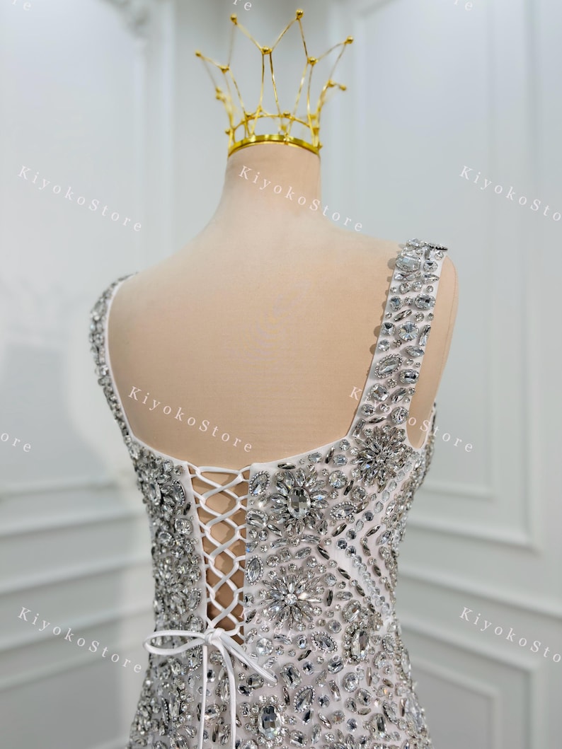 K&ouml;nnte beinhalten: Wei&szlig;es, verziertes Abendkleid mit glitzernden Kristall- und Strassdetails. Der R&uuml;cken hat einen Verschluss im Korsettstil und eine zarte Schleife. Eine goldene Krone sitzt auf einem Schaufensterpuppenkopf, was auf ein Braut- oder besonderes Anlass-Thema hindeutet.