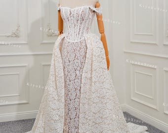 May include: A white lace wedding dress with an off-the-shoulder neckline and a fitted bodice. The dress features a long, flowing skirt with a train. The dress is displayed on a mannequin.