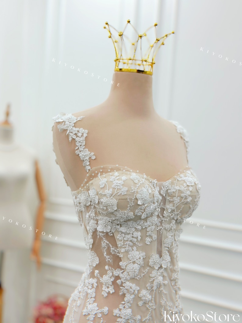 Pu&ograve; includere: Un corpetto senza maniche con una base color carne, decorato con intricati ricami floreali in pizzo bianco e piccole perle. Il capo presenta uno scollo a cuore, ampie spalline in pizzo e un design a corsetto strutturato con cuciture.