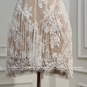 May include: A short dress featuring a nude-colored base layer with white floral lace overlays, intricate beadwork, and long, shimmering white fringe detailing that hangs from the waistline. The text KiyokoStore Custom wedding dress is visible.