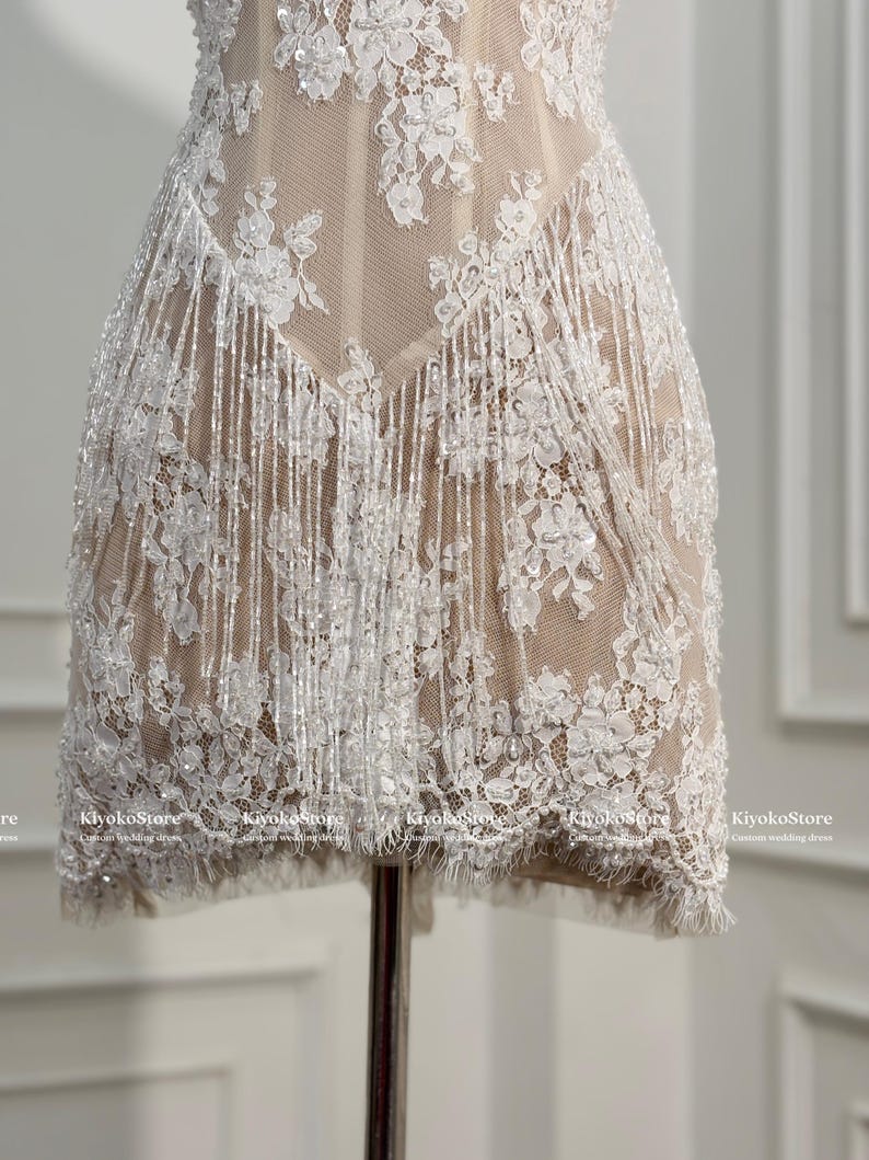 May include: A short dress featuring a nude-colored base layer with white floral lace overlays, intricate beadwork, and long, shimmering white fringe detailing that hangs from the waistline. The text KiyokoStore Custom wedding dress is visible.