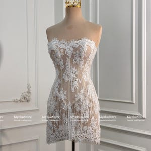 May include: A strapless mini dress featuring white floral lace embroidery over a nude-colored base, accented with shimmering sequins and delicate fringe detailing along the hem. A gold-toned crown sits atop the mannequin displaying the garment.