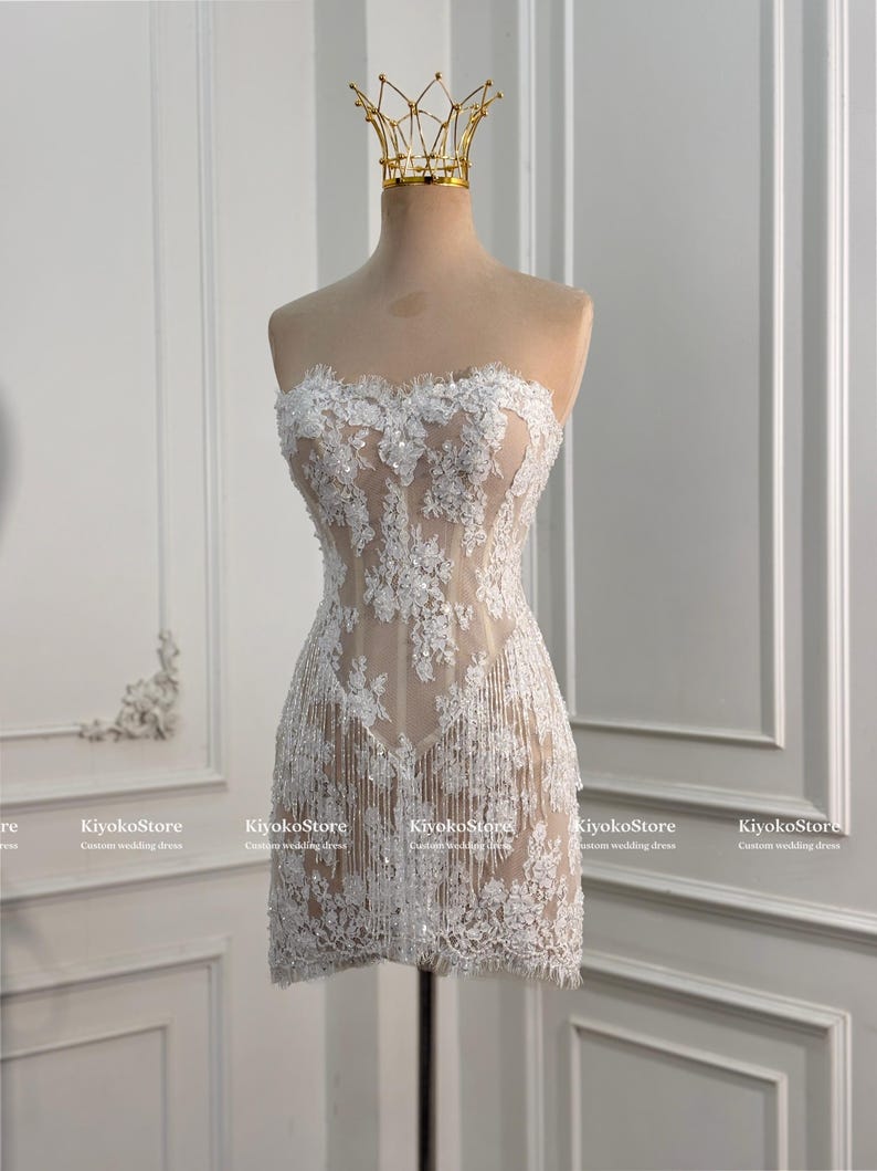 May include: A strapless mini dress featuring white floral lace embroidery over a nude-colored base, accented with shimmering sequins and delicate fringe detailing along the hem. A gold-toned crown sits atop the mannequin displaying the garment.