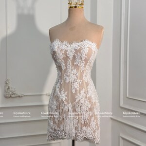 May include: A strapless mini dress featuring a nude-colored base with white floral lace overlays, intricate embroidery, and a fringe hemline. A gold-toned wire crown sits atop the mannequin displaying the dress, which includes the text KiyokoStore.